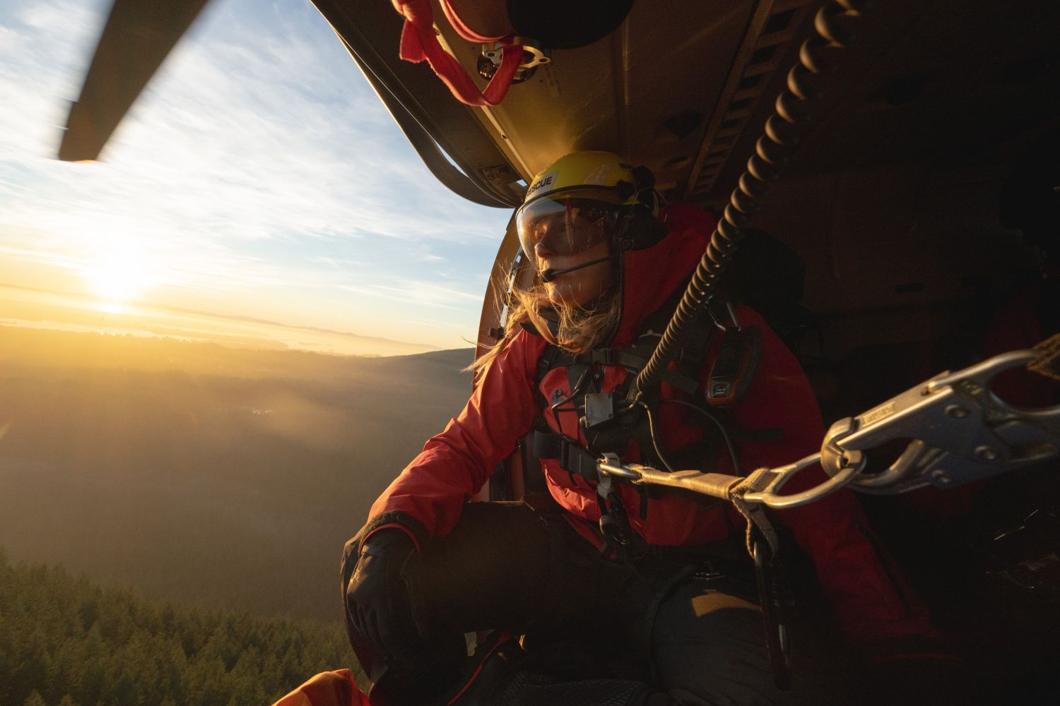 Search and rescue crew member wearing red technical Arc'Teryx PRO jacket and helmet looking out from helicopter over mountainous terrain at sunset